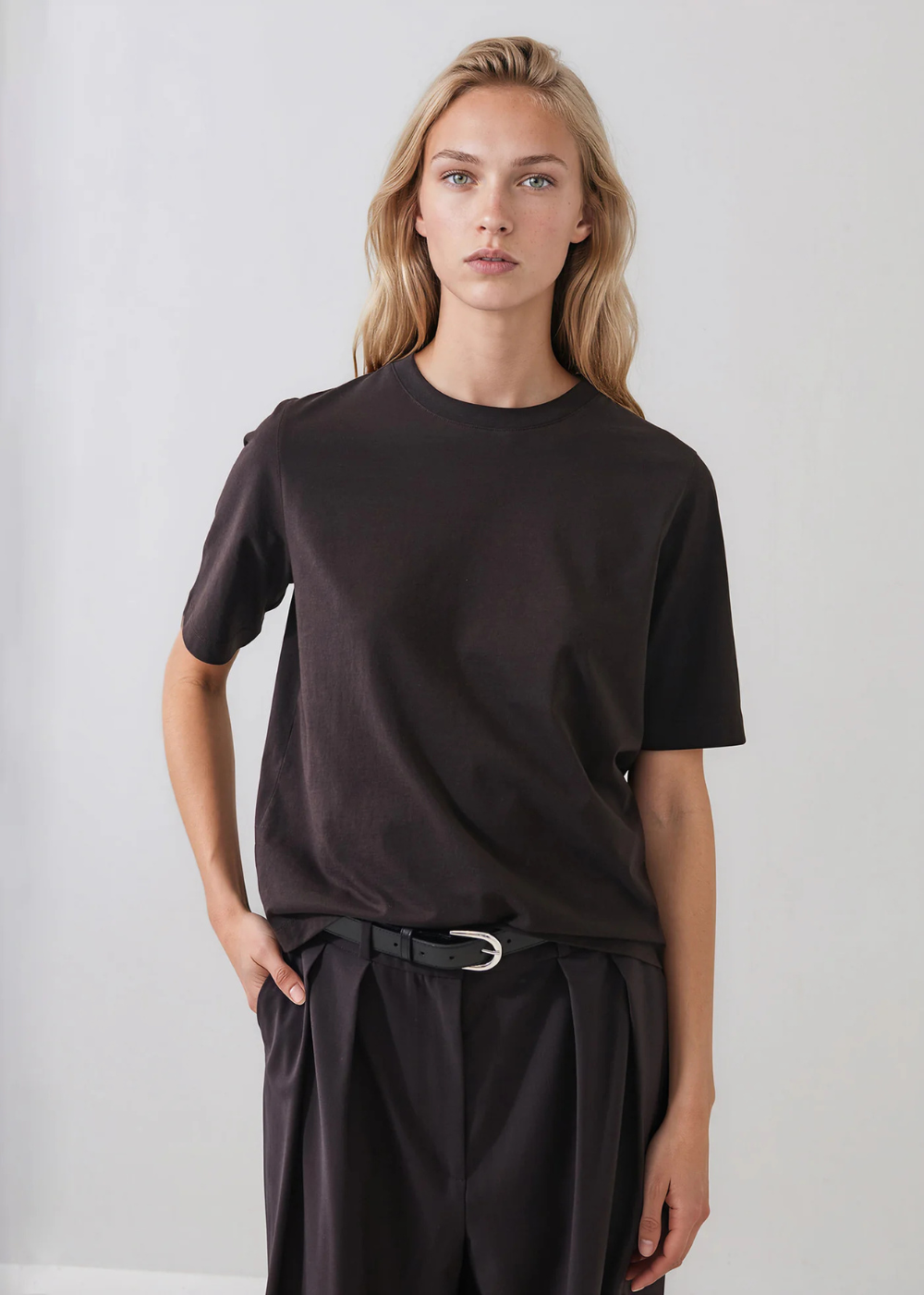 A blonde woman modeling a dark brown short-sleeve crewneck t-shirt with dark pleated trousers and a black belt, standing against a light gray studio background.
