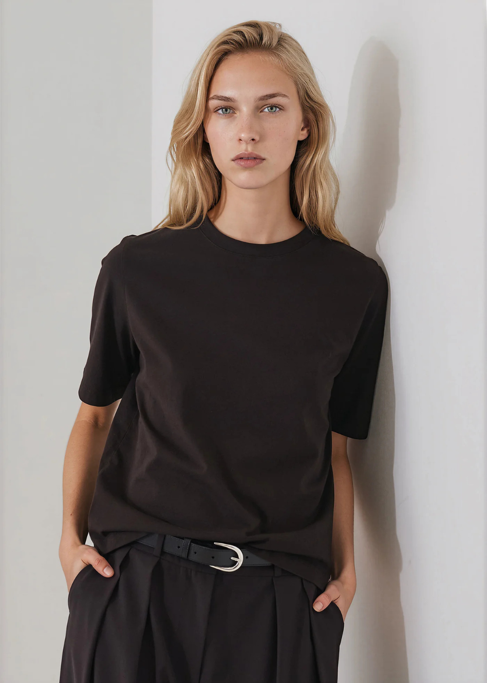 A blonde woman wearing a plain black short-sleeve crewneck t-shirt and black trousers with a black belt, standing against a light gray wall.
