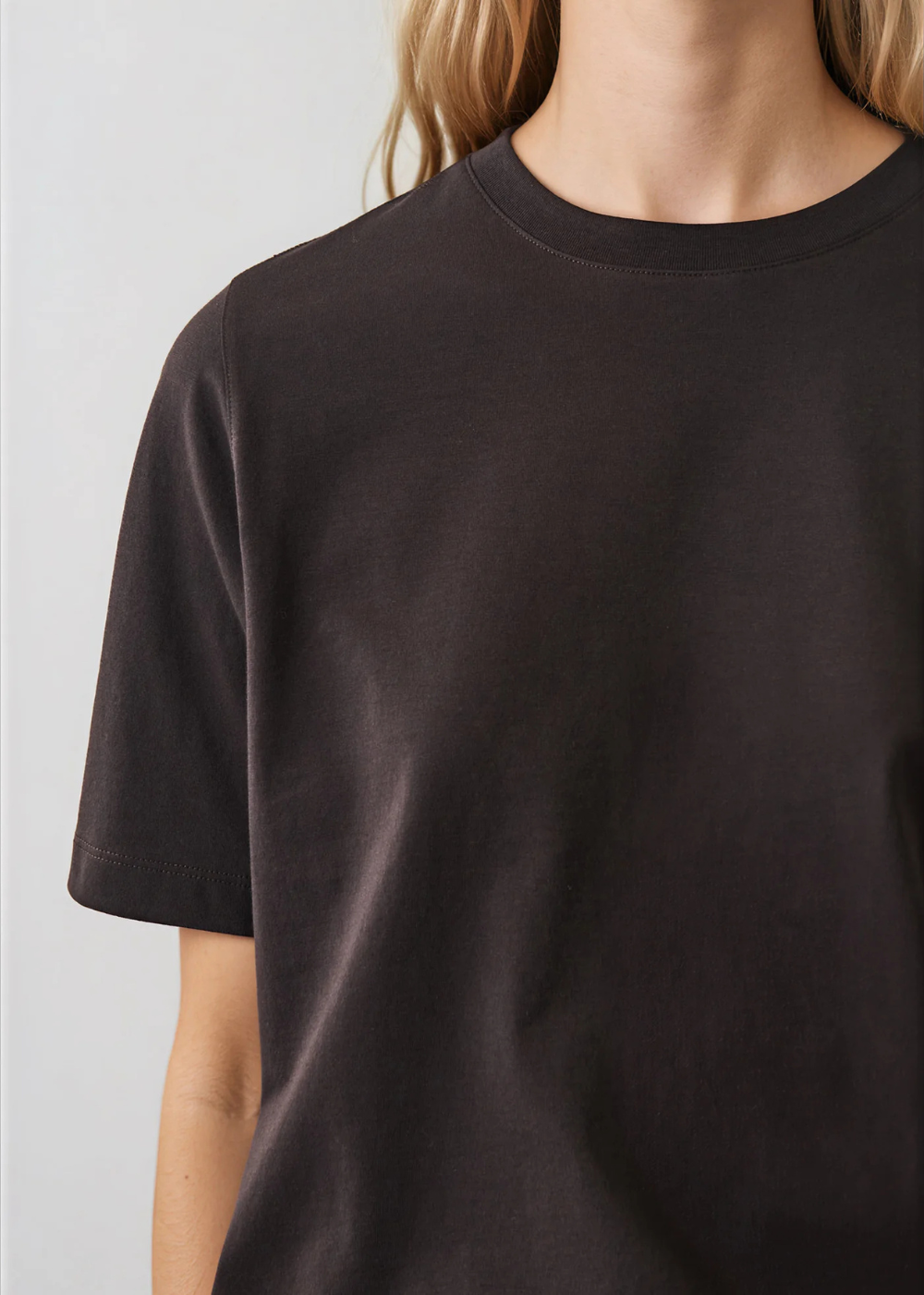 A close-up of a person wearing a plain short-sleeve crew-neck t-shirt in a very dark brown or espresso color, shown against a light gray background.