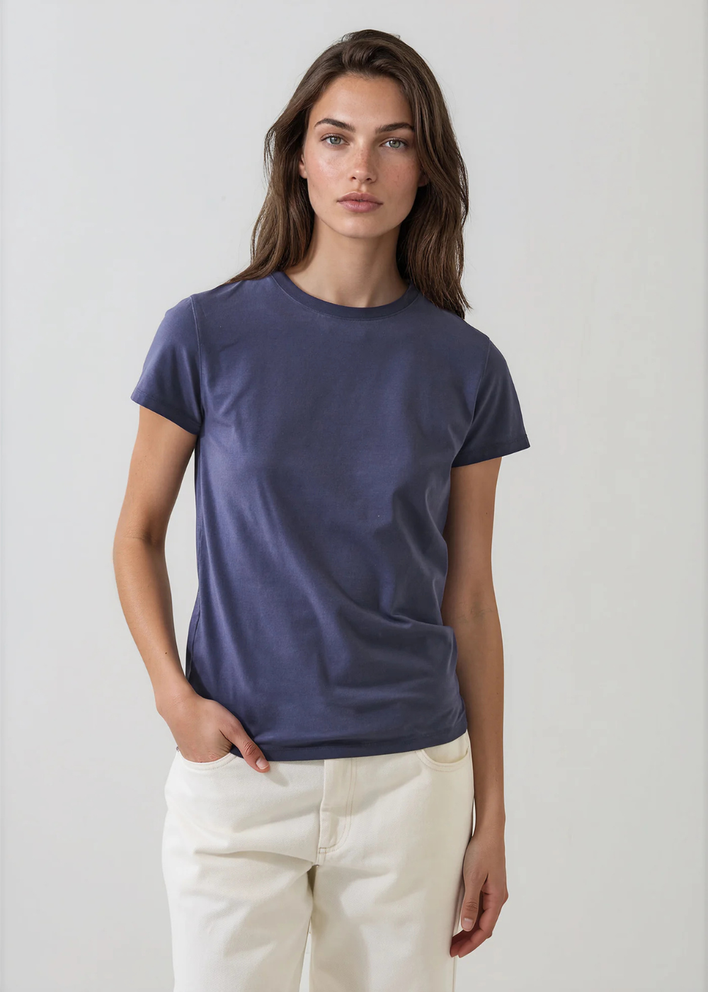 A woman modeling a short-sleeve crewneck t-shirt in a muted slate blue color, paired with off-white trousers, against a plain light gray background.