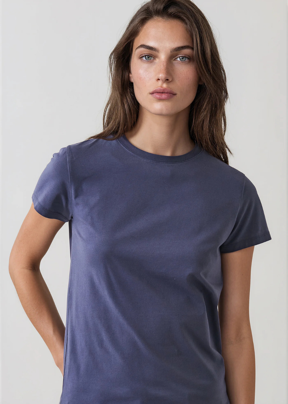 A woman wearing a plain short-sleeve crewneck t-shirt in a muted slate blue color, photographed against a light gray studio background.