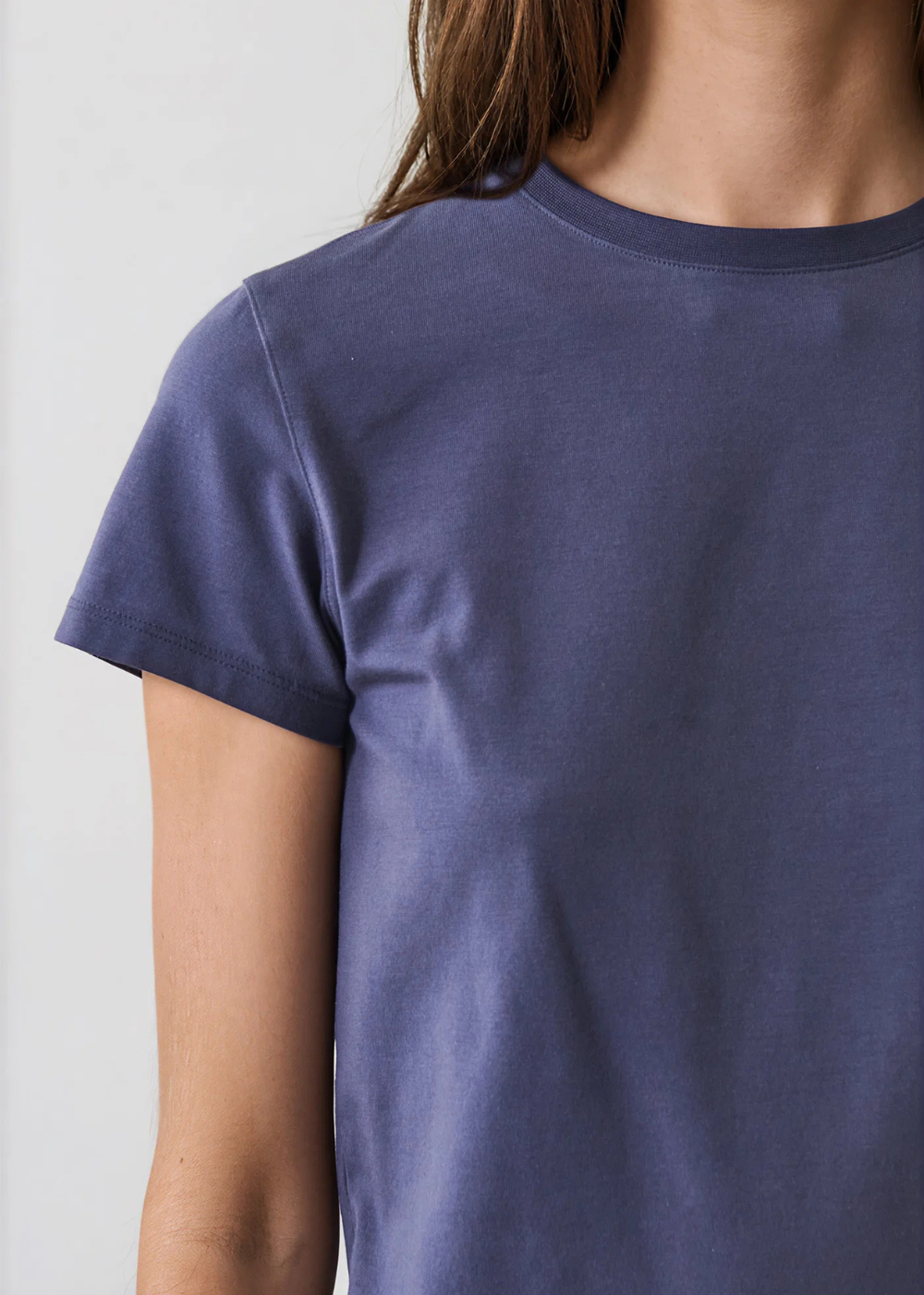 A close-up of a person wearing a short-sleeve crew-neck t-shirt in a muted slate blue color, shown from the chest up against a light gray background.