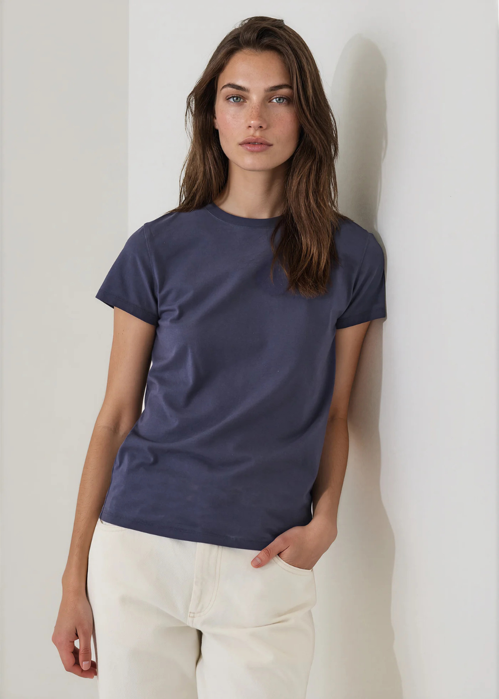A woman wearing a short-sleeve crewneck t-shirt in a muted slate blue color, paired with off-white trousers, standing against a plain light gray wall.
